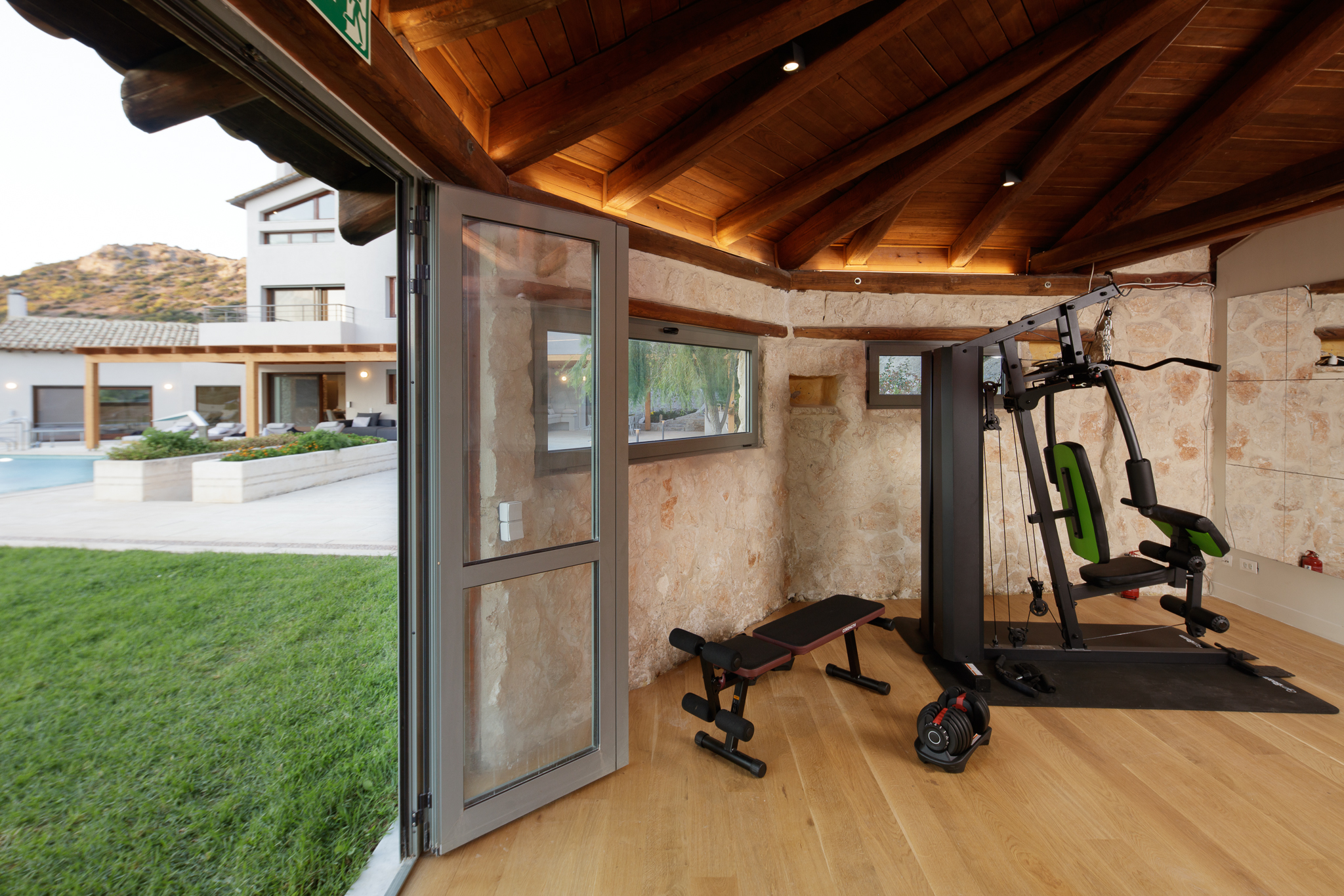 Private gym pavilion at Villa Lithos Porto Rafti with cable machine, bench and folding doors open to the lawn and pool