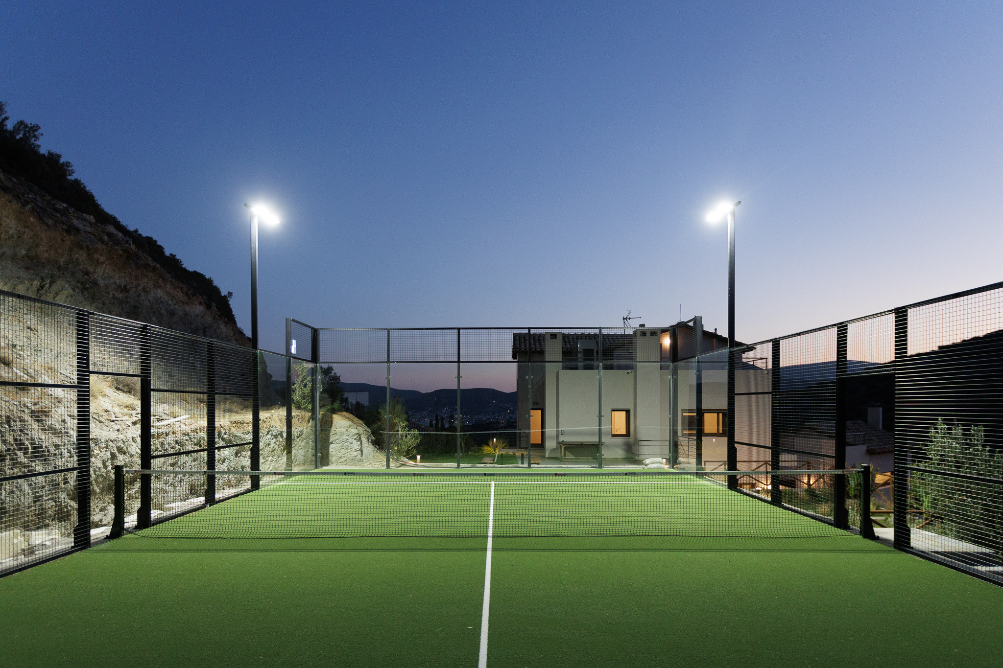 Floodlit glass-walled padel court at Villa Lithos Porto Rafti in the evening